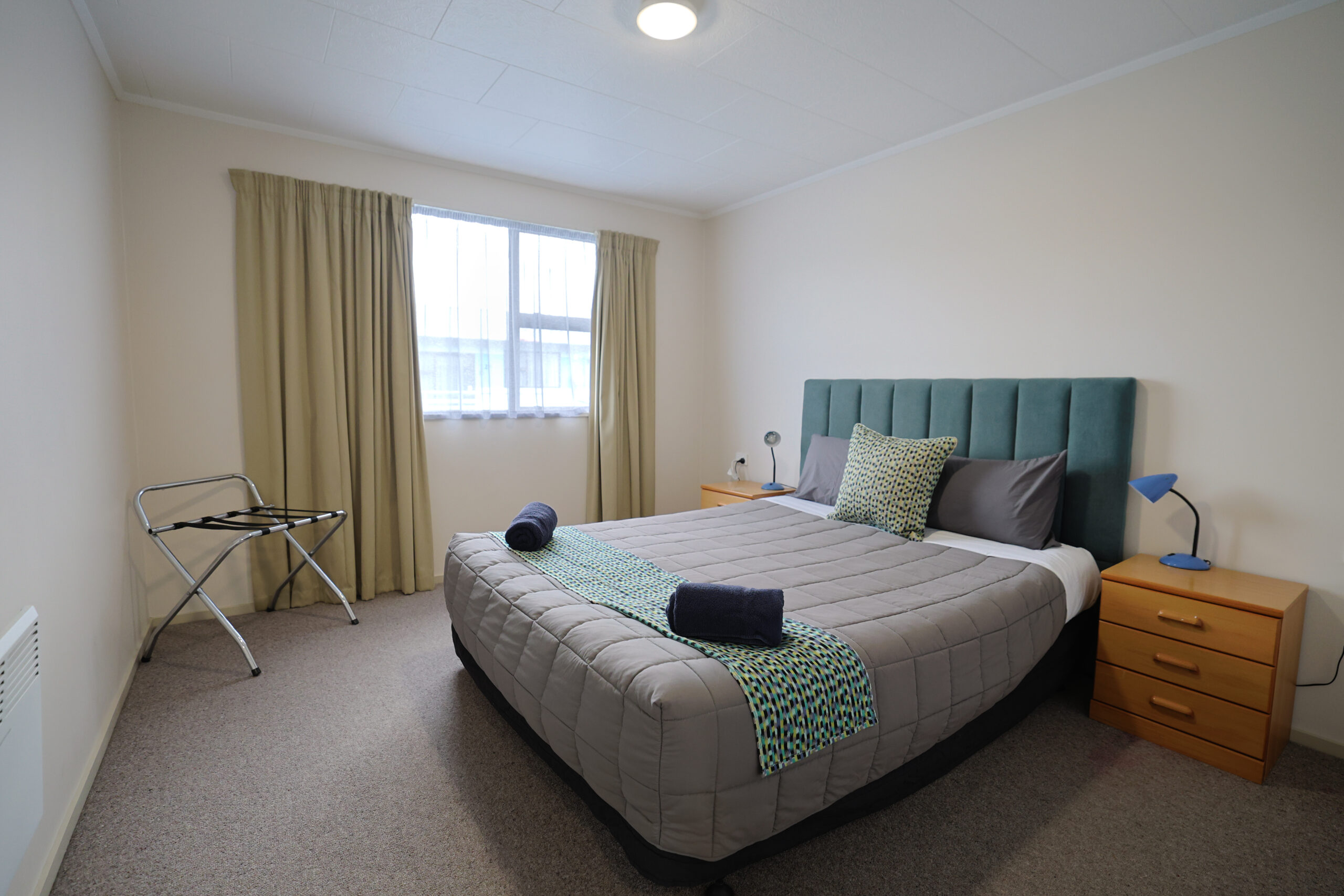 Bedroom with a teal headboard, luggage rack and natural light from a side window